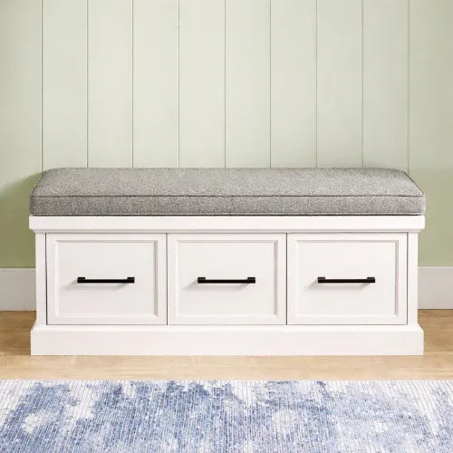 A white storage bench with three drawers and a gray upholstered seat cushion, set against a light green paneled wall with a blue rug on the floor. - view 3