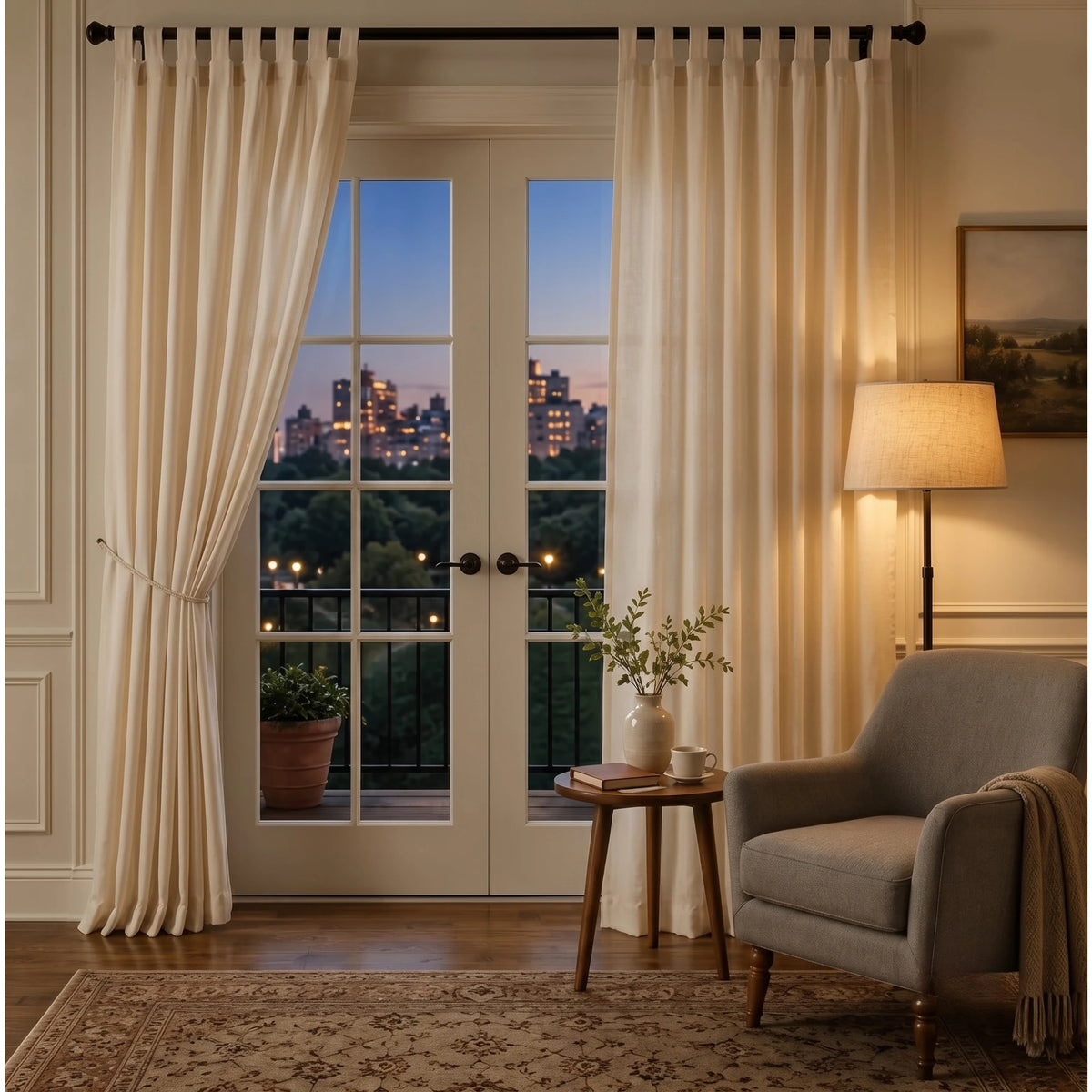 A pair of elegant cream-colored curtain panels hanging on a black metal rod in a beautifully styled room. The room features a gray armchair, a wooden side table with a vase, a lit floor lamp, and a patterned rug, with a view of a city skyline at dusk through the French doors. - view 2