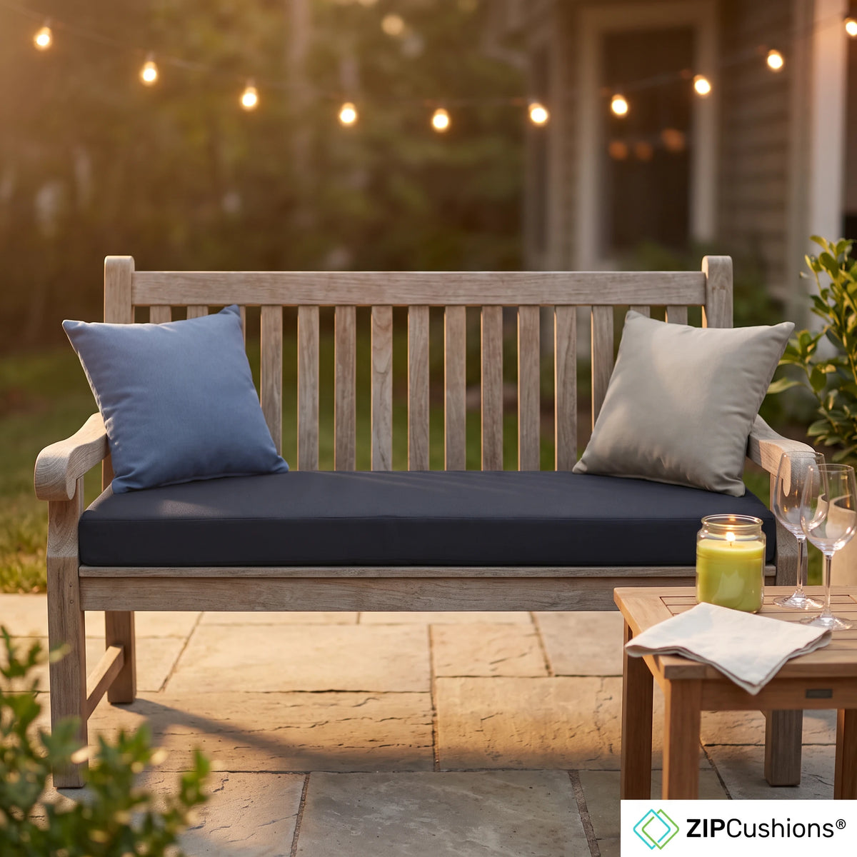 A wooden outdoor bench with thick dark blue seat cushions and two pillows, set on a stone patio at dusk with string lights and a small side table nearby. A ZIP Cushions logo appears in the bottom right corner. - view 4
