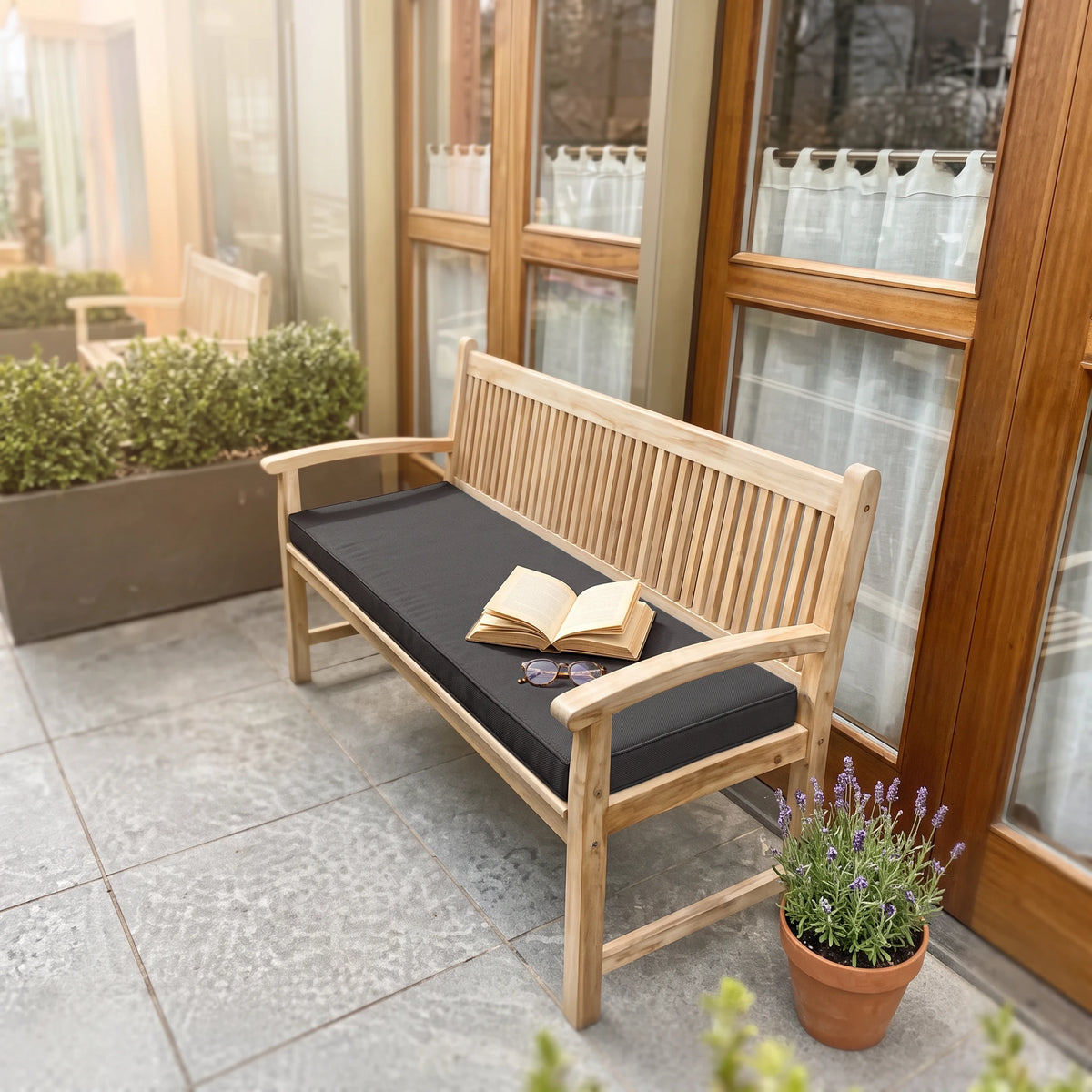 A wooden outdoor bench with a black cushion sits on a tiled patio in front of glass doors, with an open book resting on the cushion. A potted lavender plant is nearby, and the background shows a garden and house exterior. - view 3