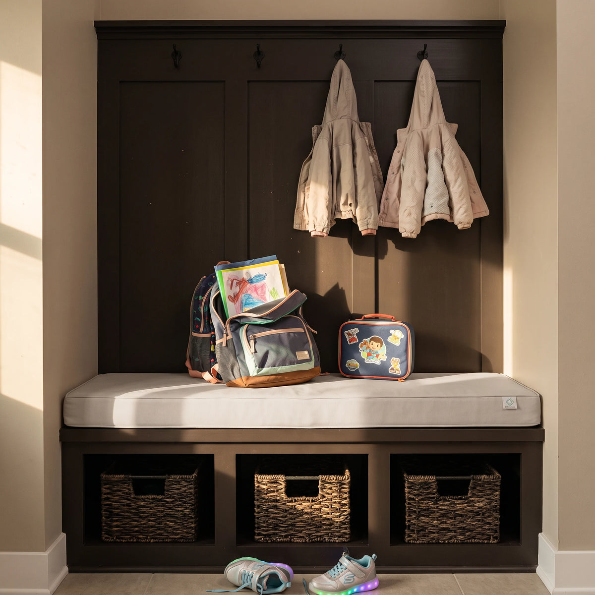 A dark wood entryway bench with a white cushion, three woven storage baskets underneath, and two hanging jackets on hooks above. A backpack, lunchbox, and books sit on the bench, with sneakers on the floor in front. - view 5