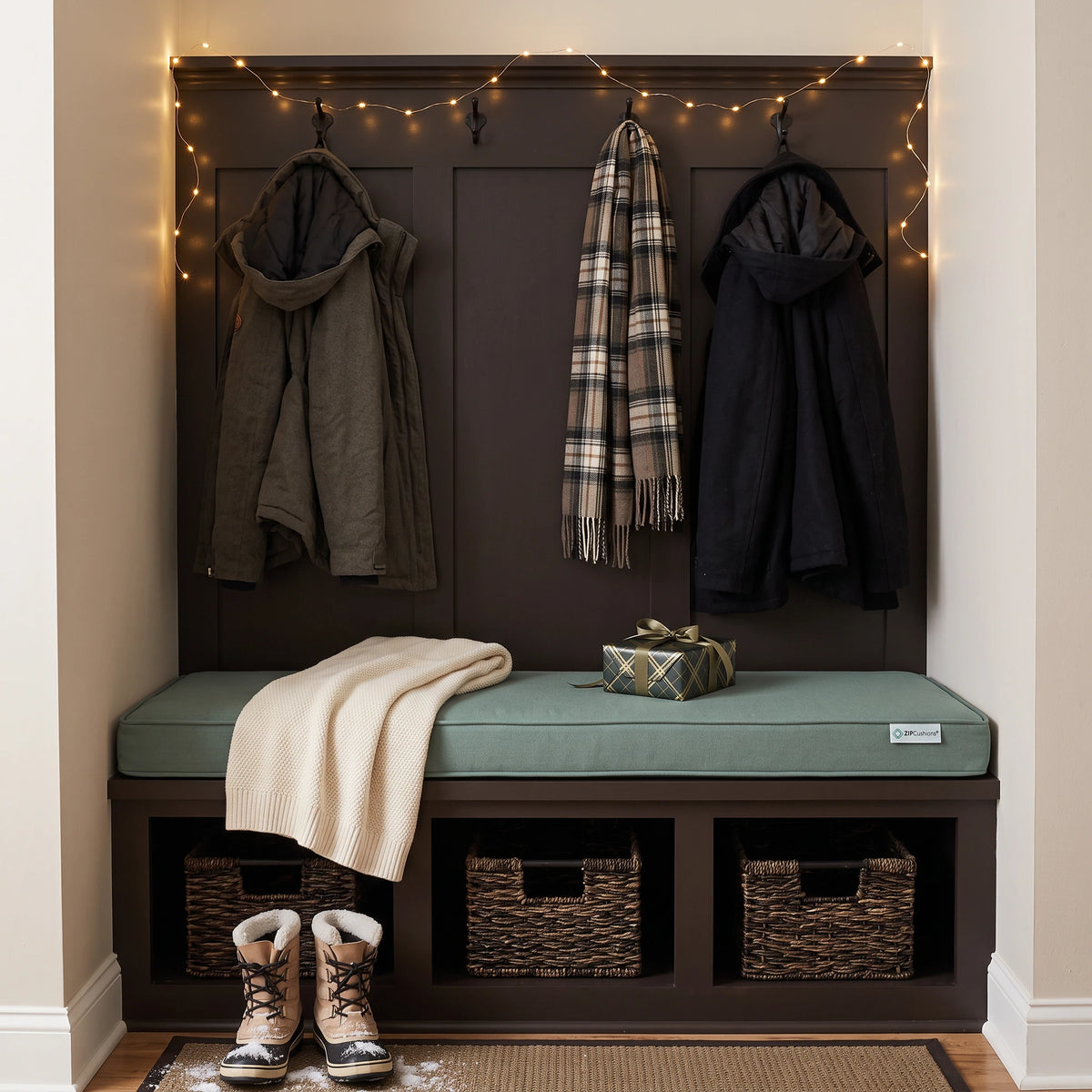 A dark brown entryway bench with a sage green cushion, three storage baskets underneath, a cream throw blanket, and a wrapped gift box on the seat. Above the bench is a matching dark brown wall panel with three hanging jackets and a plaid scarf, decorated with warm string lights.